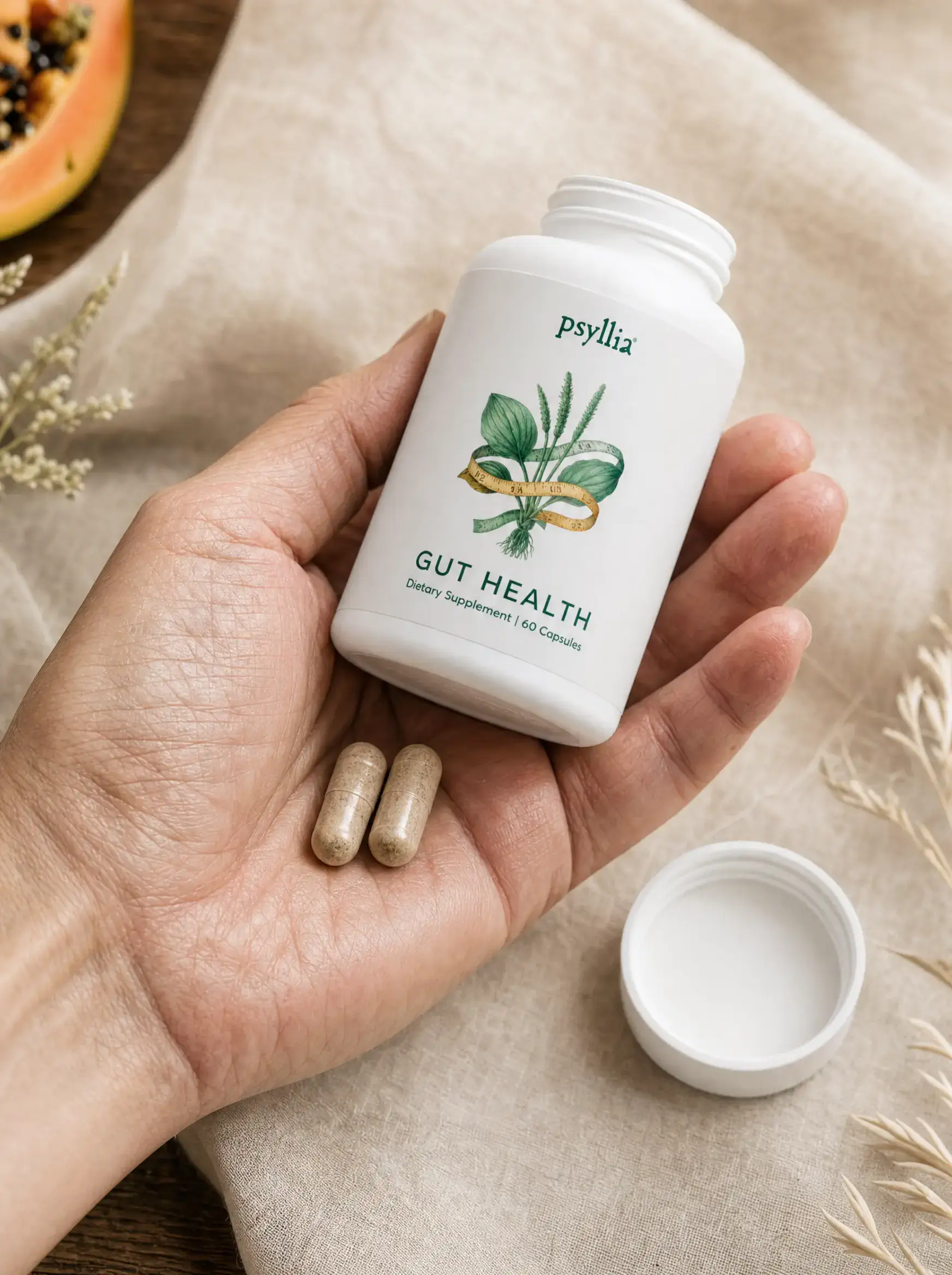 A hand holding the open Psyllia bottle with two capsules resting in the palm. The white cap is set down on warm linen, with papaya and dried grass in soft focus.